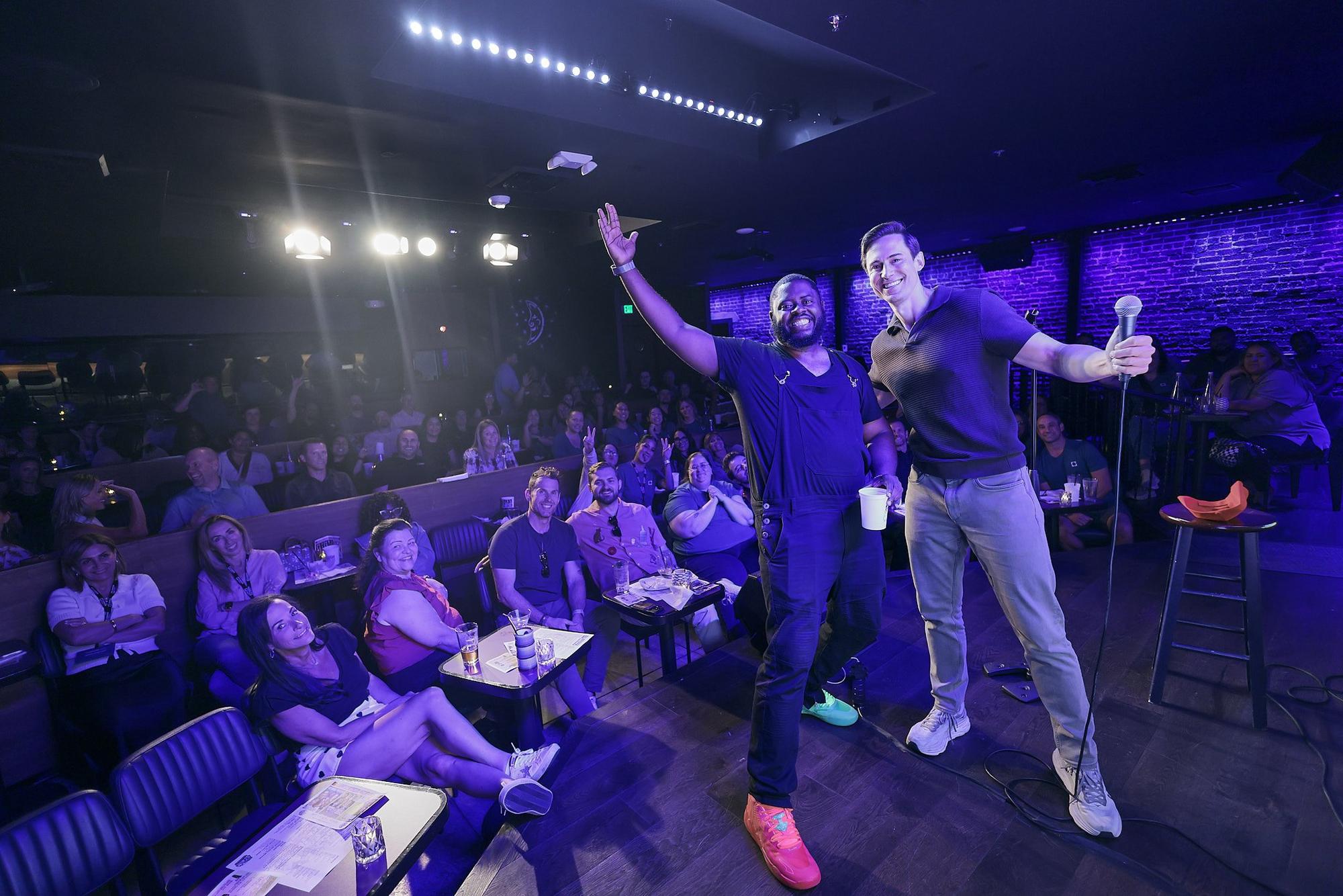 Two comics on the Ice House stage with a full corporate crowd behind them.