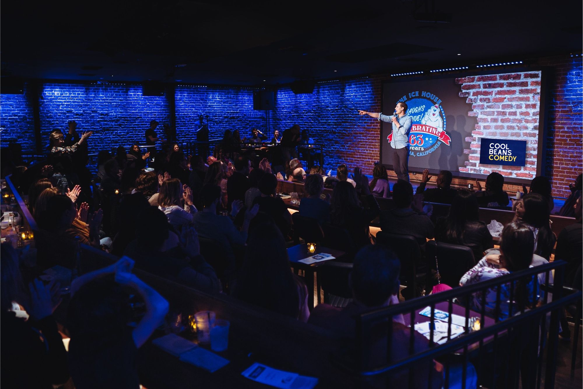 A packed, blue-lit room at the Ice House, a comic on stage pointing at the crowd, the Cool Beans Comedy logo on the screen.